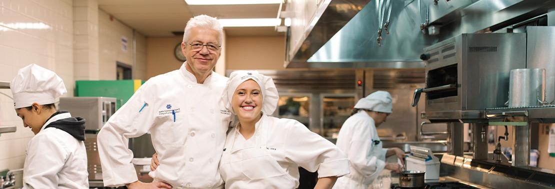 A woman and a man stand smiling in a culinary kitchen wearing culinary uniforms