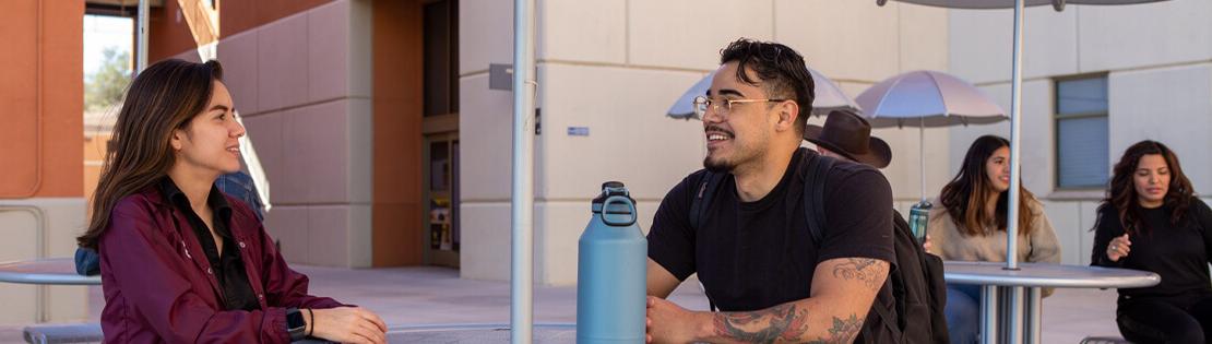 Two pima students chat at an outside table in  a courtyard at Pima's Northwest Campus